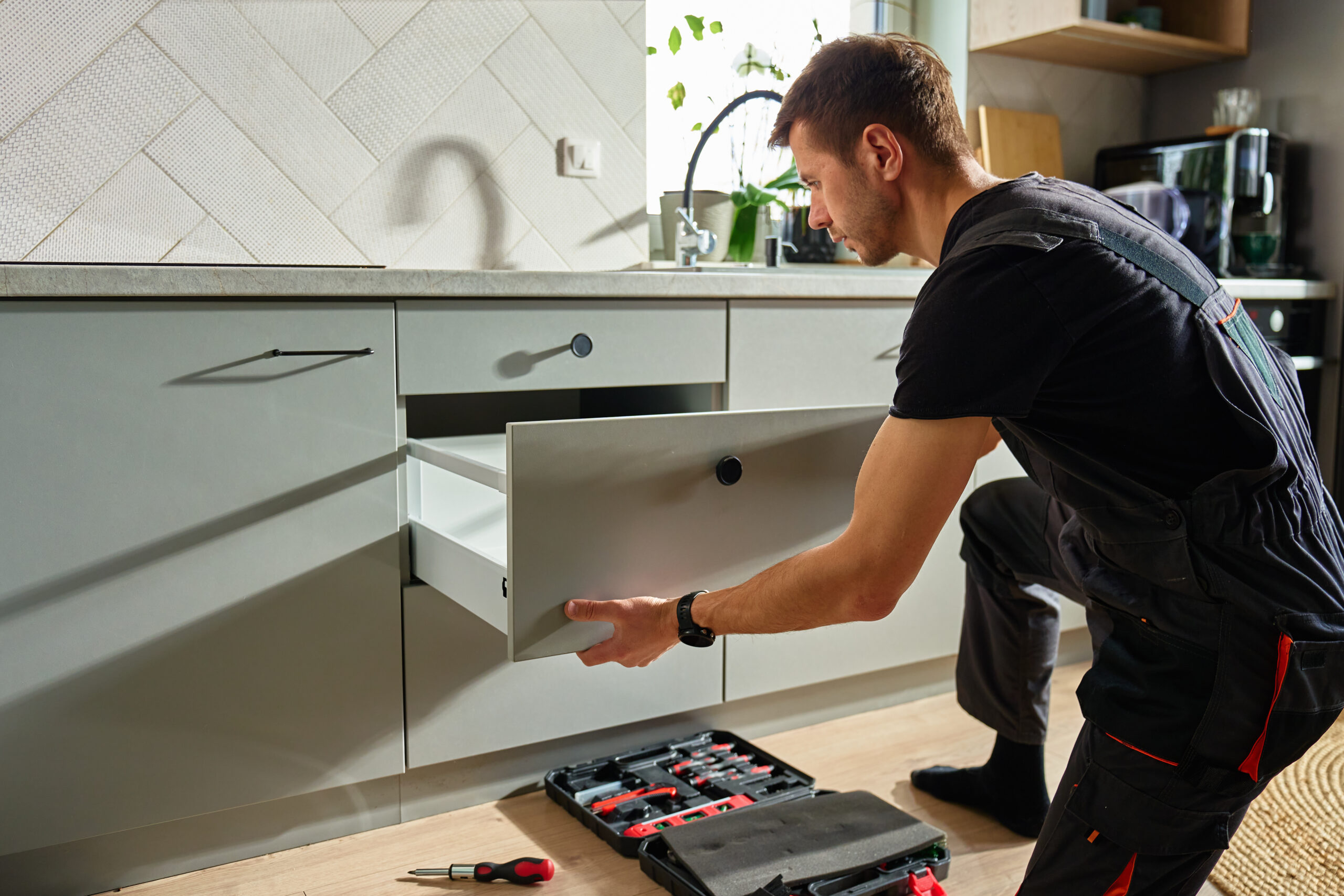 Man assembling kitchen drawer in modern cabinet using screwdriver. Handyman installing and adjusting drawer slide mechanism in contemporary kitchen. Furniture installation